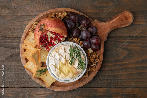 Board with tasty baked camembert, croutons, grapes, walnuts and pomegranate on wooden table, top view