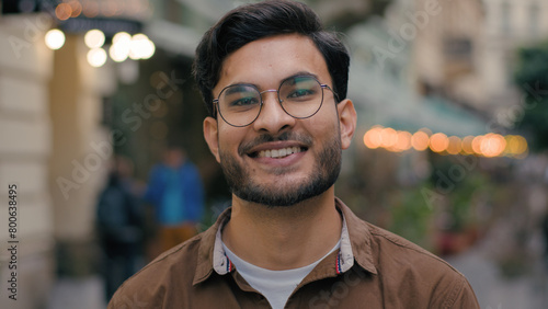 Close up portrait happy wide toothy smiling successful young handsome Indian Arabian Latino ethnic male student man guy businessman glasses posing looking friendly at camera outdoors city urban street photo