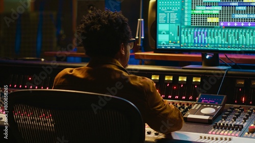 Audio expert counts down for artist singing in vocal booth, recording tracks to edit them after in post production. Producer operates mixing console and buttons or sliders, control room. Camera A.