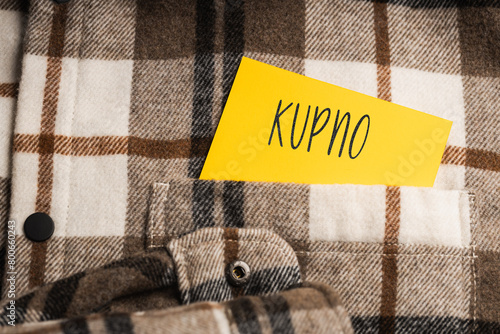  Yellow card with a handwritten inscription "Kupno", protruding from a brown plaid shirt (selective focus), translation: purchase