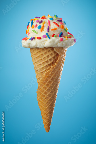 Single ice cream cone with vanilla icecream and a waffle cone covered in colorful sprinkles on blue background.
