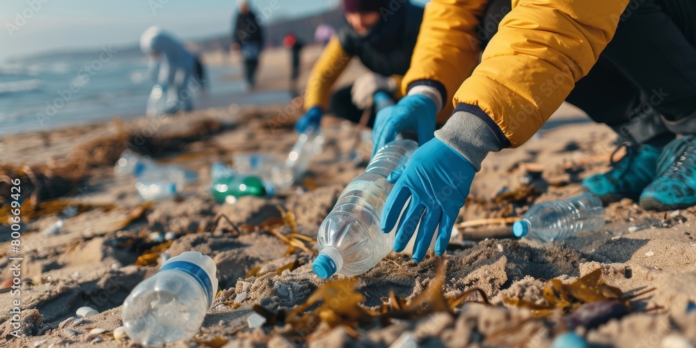 Obraz premium A person wearing gloves is picking up plastic bottles from the beach, with other people in the background cleaning and helping to clean it up.