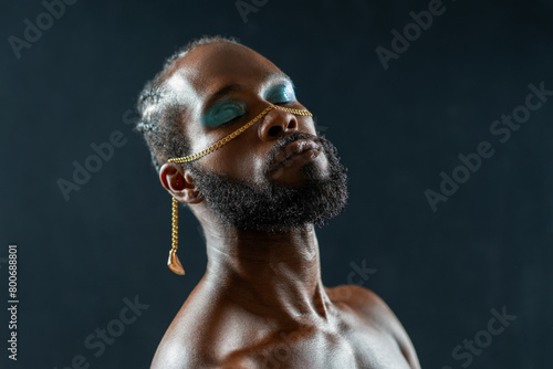 Portrait of shirtless African American gay man with blue eyeshadows and golden chain on face with closed eyes against dark blue background