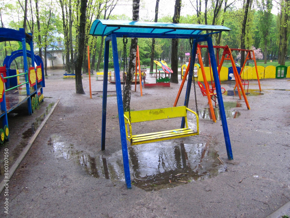 Empty swing seat in public park outdoor kid playground, wet rainy day, rain and puddles. Empty swing seat in public park outdoor kid playground, wet rainy day, rain and puddles.