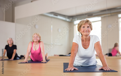 Group of aged ladies doing cobra pose on rugs in fitness room.
