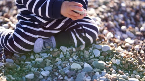Small baby in a striped overalls sorts through the pebbles while sitting on the beach. High quality 4k footage photo