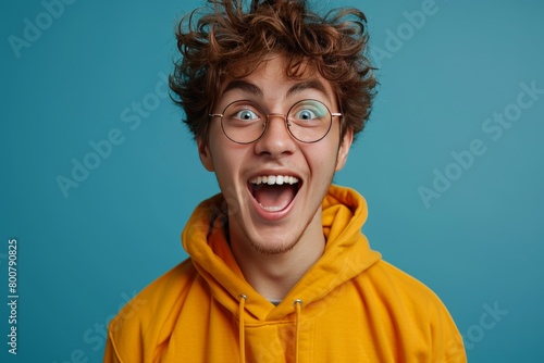 Excited young man in yellow hoodie and glasses looking at camera isolated over blue background © Rasheeda