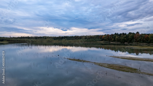 The image conveys a moment of tranquility at dusk with a view across a calm lake reflecting the evening sky. The horizon is lined with a forest showing early signs of autumnal color  adding a touch of