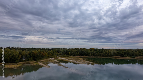 The photograph presents a vast landscape where a still lake reflects the complex patterns of a brooding sky  heavy with clouds. The surrounding forest  a lush canvas of greens  anchors the scene