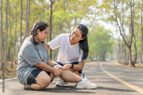 Overweight woman with knee injury sitting on the running track at a running park holding her painful knee while her female running friend sitting beside her in concern