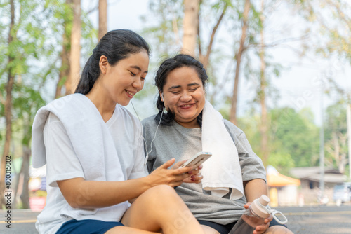 Two happy Asian female friends listening to music from a phone while sitting together at the curveside road after their morning run