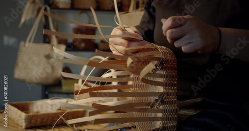 Slow motion scene while a beautiful Asian female artisan who is a local villager weaving folk basket made from leftover paper band to sell as extra income in the basketry shop shop.