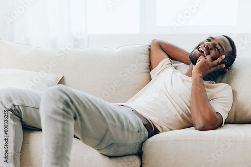 Happy African American man sitting on a black sofa, holding a smartphone and engaged in a video call He is dressed casually in a white tshirt and is comfortably relaxing in his modern apartment The