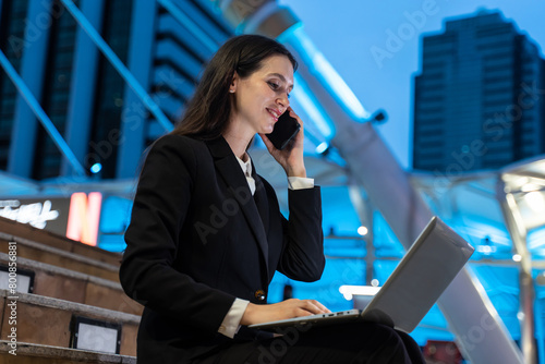 Caucasian businesswoman using laptop and phone working in city at night. 
