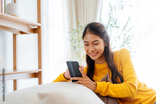 Happy young Asian woman sitting on sofa holding mobile phone using cellphone technology doing ecommerce shopping, buying online, texting messages relaxing on couch in cozy living room at home