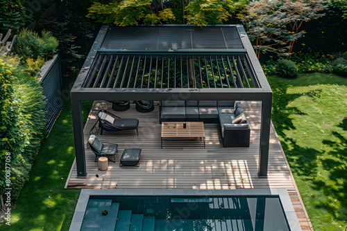 Top view of a modern black bio climatic pergola over an outdoor patio with a pool, teak wood flooring, lounge chairs, and lush green garden.