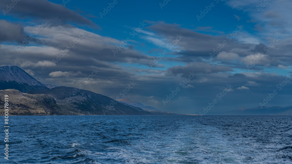 The foam trail behind the stern of the ship stretched across the surface of the blue endless ocean. Ripples on the water. Mountains against the sky and clouds. Beagle Channel.  Argentina.