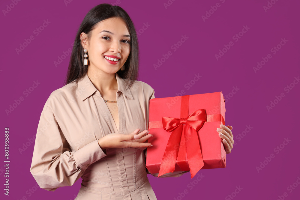 Beautiful young Asian woman pointing at gift box on purple background Beautiful young Asian woman pointing at gift box on purple background