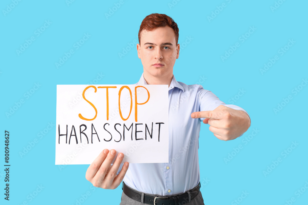 Young businessman pointing at paper with text STOP HARASSMENT on blue background Young businessman pointing at paper with text STOP HARASSMENT on blue background