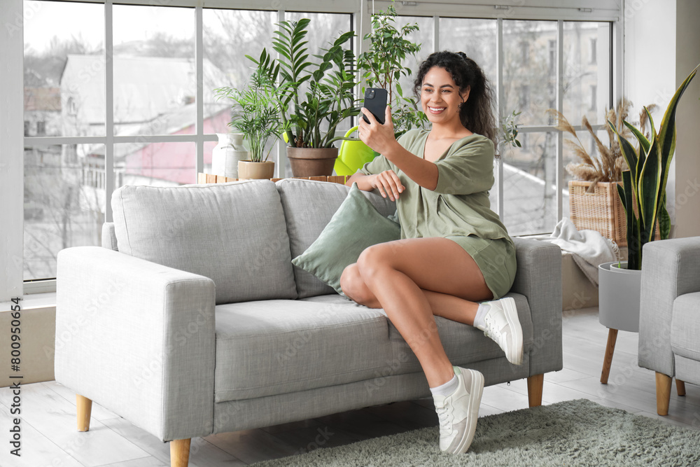 Fototapeta premium Beautiful young African-American woman taking selfie with plants in living room