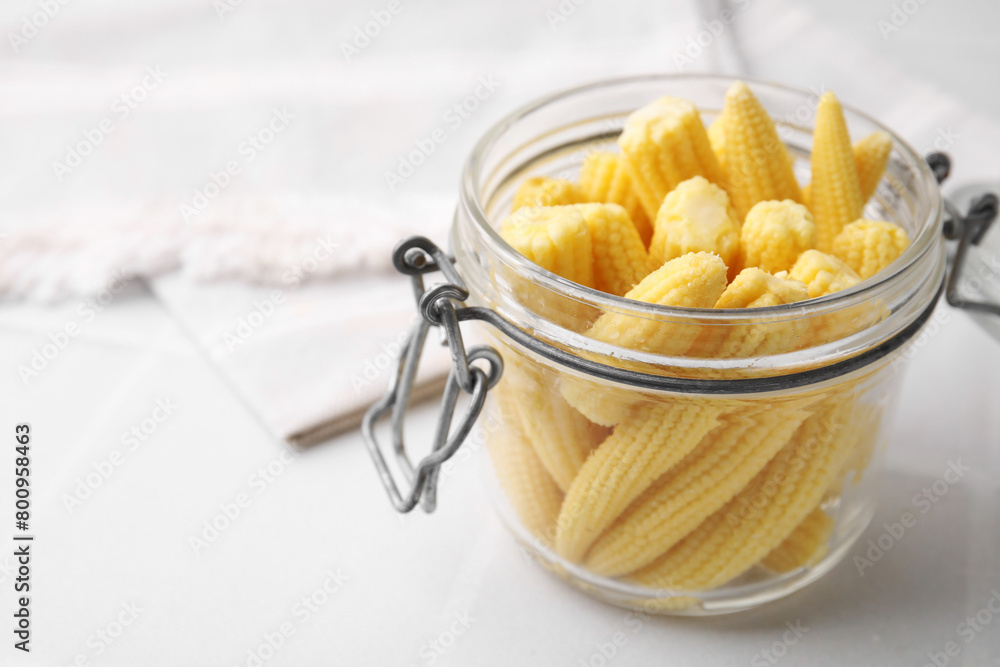 Tasty fresh yellow baby corns in glass jar on white tiled table, closeup. Space for text
