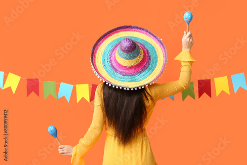 Young woman in Mexican sombrero hat, with maracas and festive garland on orange background, back view