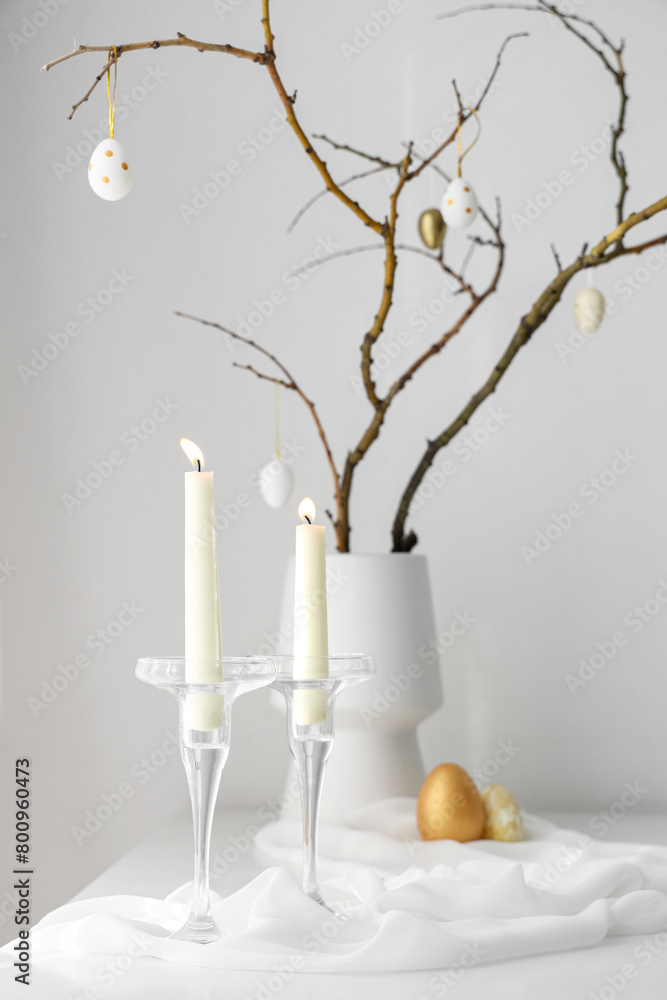 Burning candles, vase with tree branches and Easter eggs on table near light wall, closeup