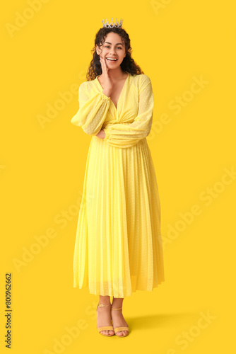Happy young African-American woman in stylish prom dress and tiara on yellow background