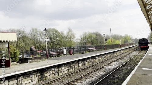 A slow motion video of an old fashoned steam train arrives from the distance and begins to slow as it approaches the station platform, on a cloudy day in the United Kingdom. photo