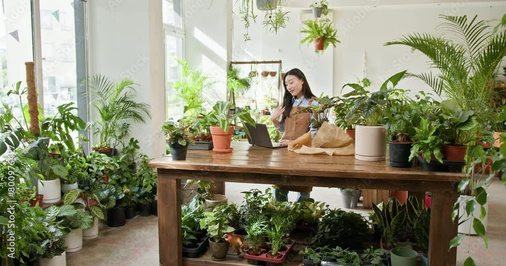 Smiling Asian flower shop owner working on laptop computer. checking stocks, taking customer orders, selling products online. Occupation working concept. Flower shop. Small business involvement. Smiling Asian flower shop owner working on laptop computer. checking stocks, taking customer orders, selling products online. Occupation working concept. Flower shop. Small business involvement.