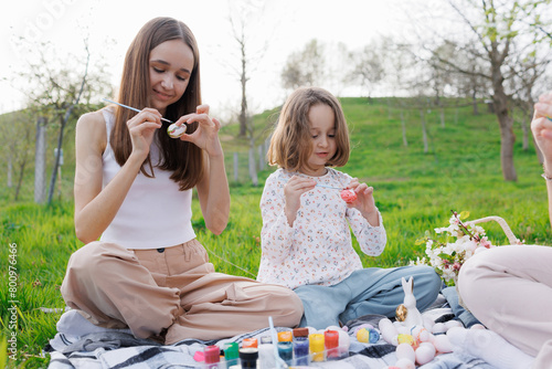 A young girl and her mother are sitting on a blanket in a park  painting Easter eggs. Scene is peaceful and relaxing