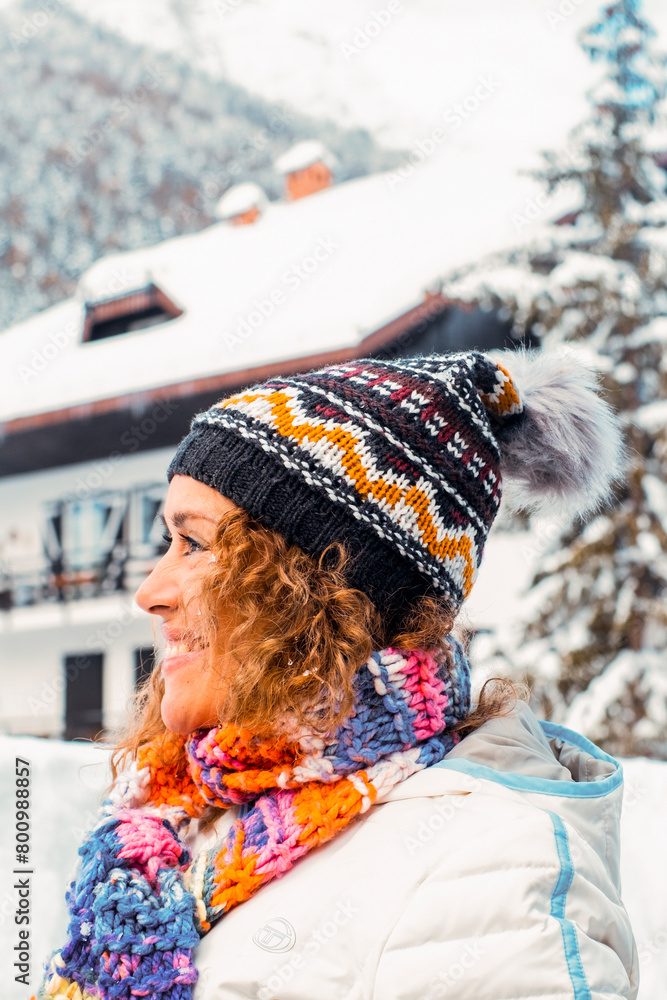 Side portrait of happy adult woman smiling and enjoying snow mountain outdoor leisure activity alone. Beautiful wood chalet house in background with trees pine in the garden outside. Winter holiday