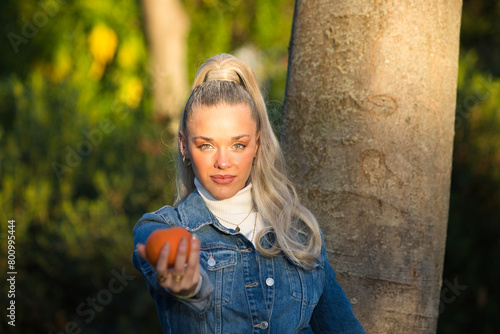 Pretty young blonde woman with a ponytail in her hair wearing a denim dress holds an orange in her hand and holds it out in front of her. Focus on the girl and the orange out of focus. 