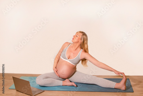 Health concept. Young beautiful pregnant woman does yoga exercise in the modern room. A woman does yoga at home online on a laptop. Against the background of a white wall.