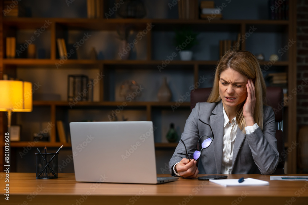 Businesswoman sitting alone in her home office and suffering from a headache. Young businesswoman experiencing fatigue during her work day at home. Businesswoman sitting alone in her home office and suffering from a headache. Young businesswoman experiencing fatigue during her work day at home.