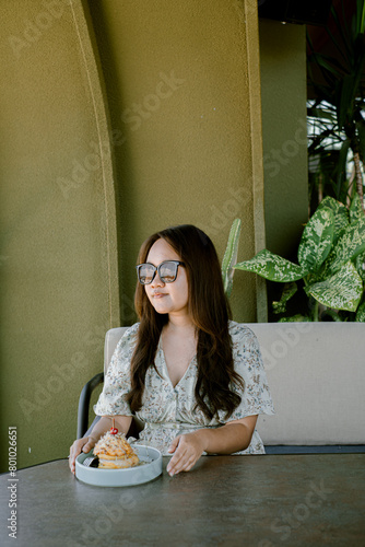 An Asian woman sits and eats a cake in the semi-outdoor dining area. The woman was wearing a tropical dress and sunglasses.
