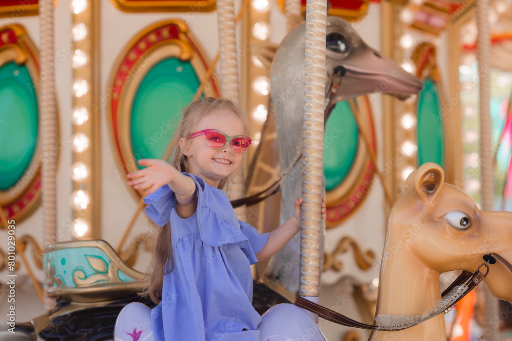 Fototapeta premium A happy little girl rides a carousel in the summer at an amusement park