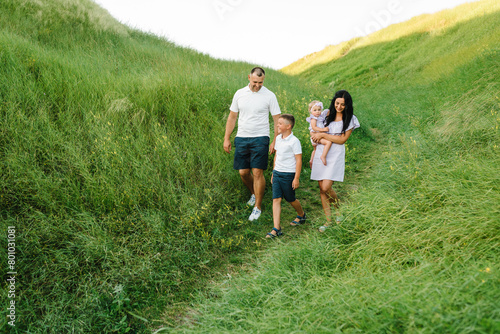 Happy young family spending time together in mountains. Mom, dad, daughter girl and son boy walk in green grass in field. Children hold hands parents in nature. Family holiday at sunset on summer day.
