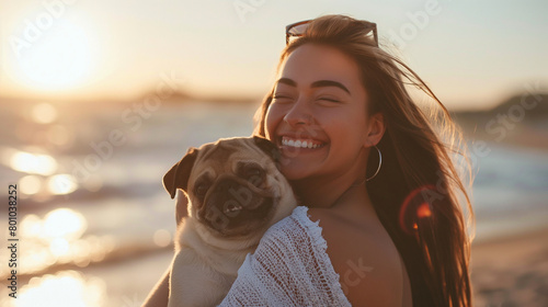 With a radiant smile, the woman cradles her pug in her arms, their bond evident in the way they share affectionate glances amidst the tranquil setting of a beach at sunset. photo