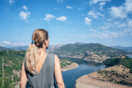 Rear view of a young long-haired woman enjoying the view of one of most picturesque meander of Arda river surounded by mountain slopes of Rhodopi Mountains, Bulgaria.