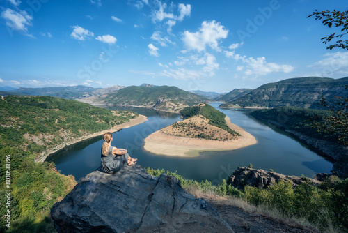 Rear view of a young long-haired woman enjoying the view of one of most picturesque meander of Arda river surounded by mountain slopes of Rhodopi Mountains, Bulgaria. photo