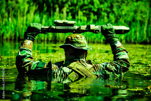 A camouflaged soldier walks through a swamp, submerging himself, his arms and rifle visible, shot from behind, realities of harsh military service
