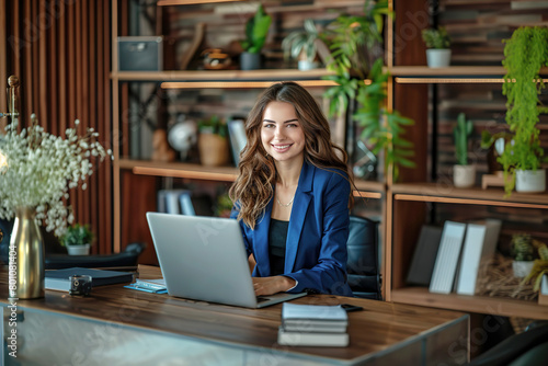 Beautiful businesswoman in her office. Smiling woman dressed in blue clothes in an office with a laptop, sitting at a table with helves in the background.