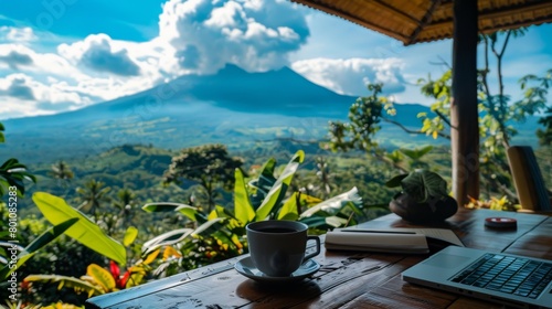 A wooden table with a cup of coffee, a notebook and a laptop on it. In the background there is a volcano and a jungle.
