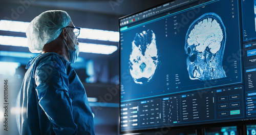 Fotografia Hospital Research Laboratory: Portrait Of Caucasian Male Neurosurgeon Looking At TV Screen With Brain MRI Scans Of Patient, Analyzing, Preparing For Complicated Neurosurgery, Wearing Surgical Gown