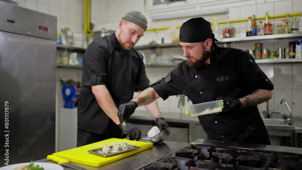 Confident male cook in a black uniform and bandana sprinkles a finished dish with cheese in the kitchen. Duo of chefs in the kitchen preparing a dish together Confident male cook in a black uniform and bandana sprinkles a finished dish with cheese in the kitchen. Duo of chefs in the kitchen preparing a dish together