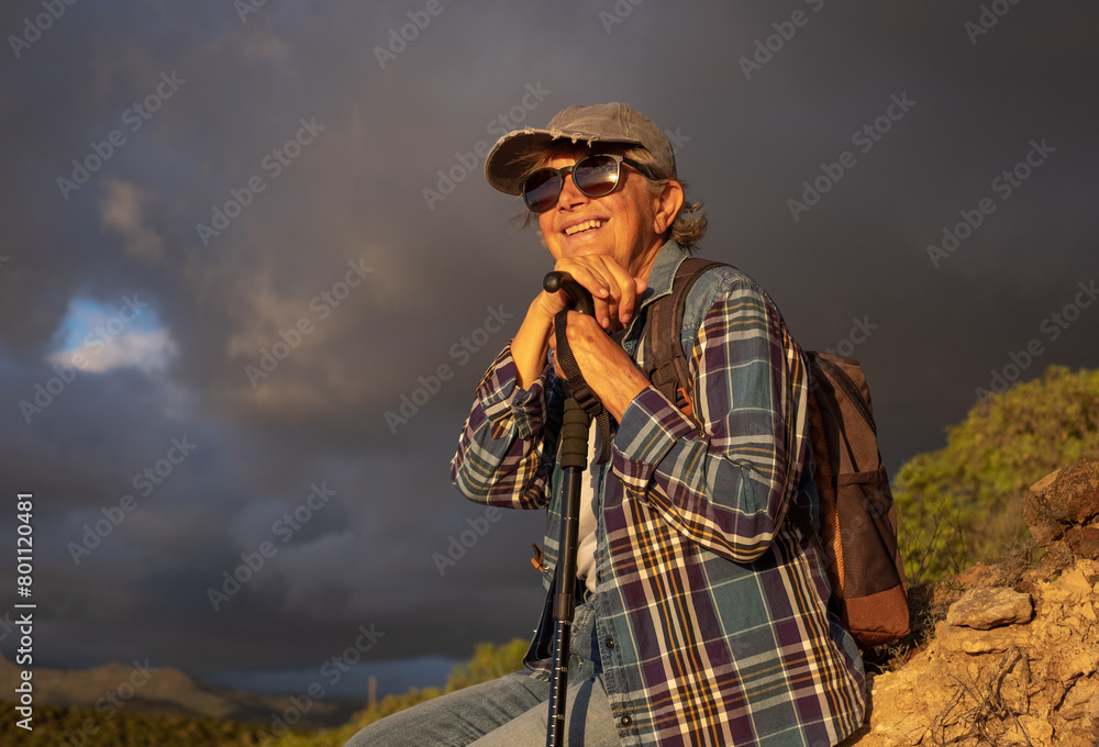 Happy senior woman with hat sitting on a hike outdoors in sunset light with dramatic sky ready to make a big thunderstorm. Healthy lifestyle, vacation and sporting activity in retirement