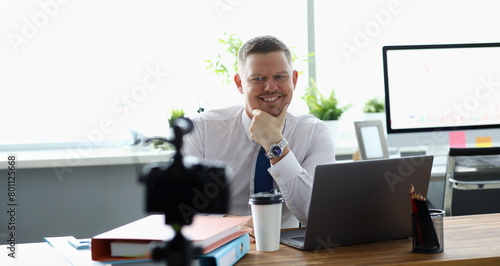 Portrait of cheerful man sitting at workplace in office. Blogger drinking coffee at workspace and making video for financial vlog. Business concept. Blurred background