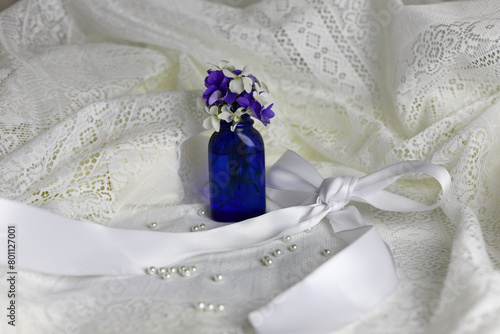 Violets in an antique blue bottle. White satin bow, pearls and lace background.