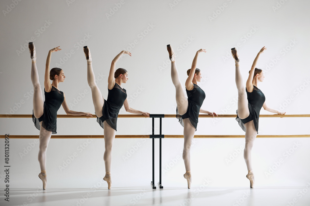 Young girls, ballet dancers showing grace and flexibility, standing next to barre and training, practicing against grey studio background. Concept of ballet, art, dance studio, classical style, youth